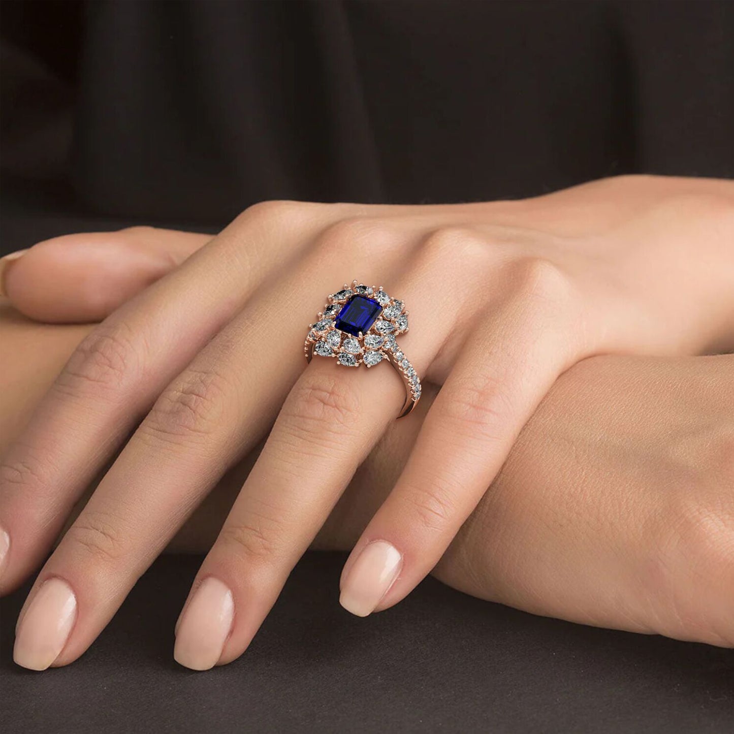 Hand wearing a 14k rose gold ring with a 2.50CT blue sapphire and halo diamonds against a dark background