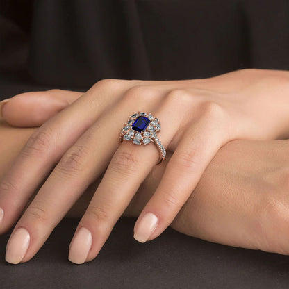 Hand wearing a 14k rose gold ring with a 2.50CT blue sapphire and halo diamonds against a dark background