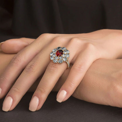 A hand adorned with a 14k gold 2.50CT oval red ruby ring surrounded by clear diamonds, resting on a dark surface, exuding elegance and luxury.