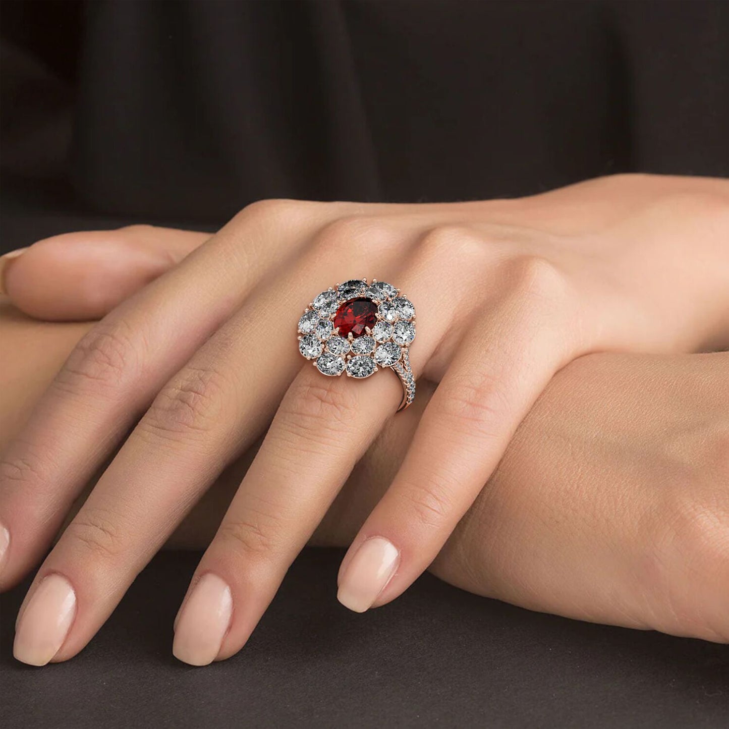 A hand wears a 14k rose gold ring with a 2.50CT oval ruby surrounded by clear diamonds, set against a soft black fabric background.