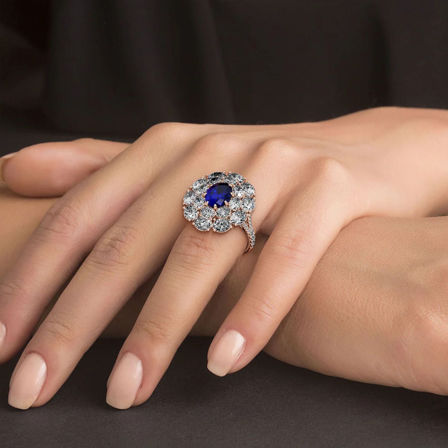 A hand elegantly displays a 14k rose gold ring with a 2.50CT oval blue sapphire surrounded by clear diamonds against a dark background.