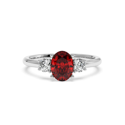14k white gold oval ruby ring with red center gemstone flanked by two round white diamonds. Elegant, luxurious design.